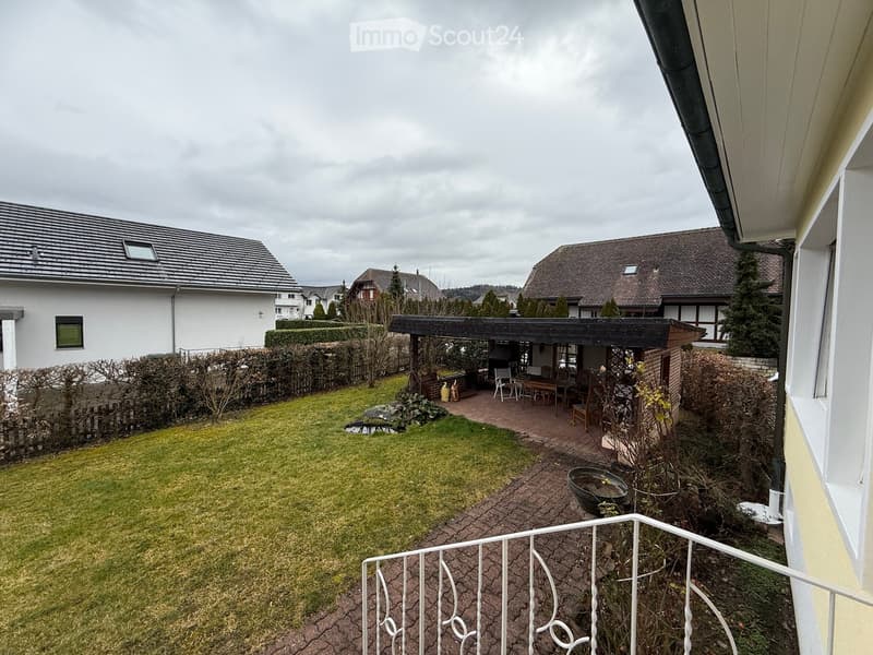 3 houses visible, fence, shrubs, garden with wooden gazebo and patio furniture, brick pathway, small pond, plants, trees, balcony