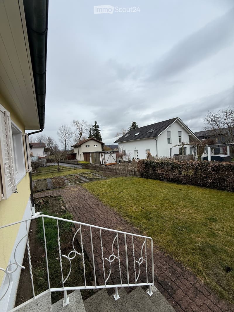 Garden with a brick pathway, white fence, green lawn, yellow building, trees, house
