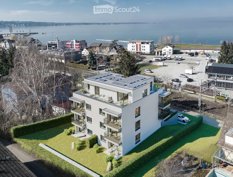 Modern white building, elevated from the ground, with solar panels, a view of the sea, multiple windows, balconies, a nearby parking lot, and green lawns.