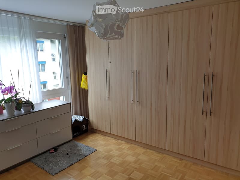 wooden wardrobes, wooden floors, white dresser, rug, window with white curtains, flowers in pots on dresser