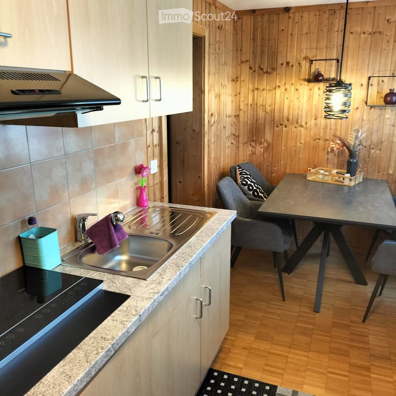 Kitchen includes a sink, stove, and cabinets. The table in the dining area has a vase and a basket with tea cups. Wooden walls are adorned with picture frames and a light fixture.