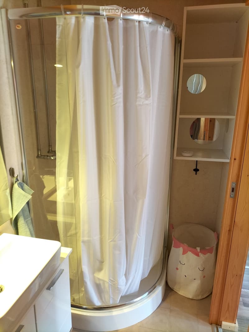 glass shower enclosure with a white curtain, white sink, white floor and wall tiles, wooden cabinet, round mirror, storage bag