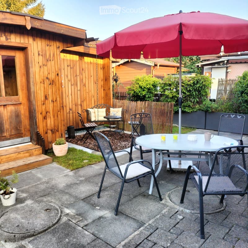 Table with chairs, wooden bench, small table with stools, red umbrella, pot plants, grass, wooden wall