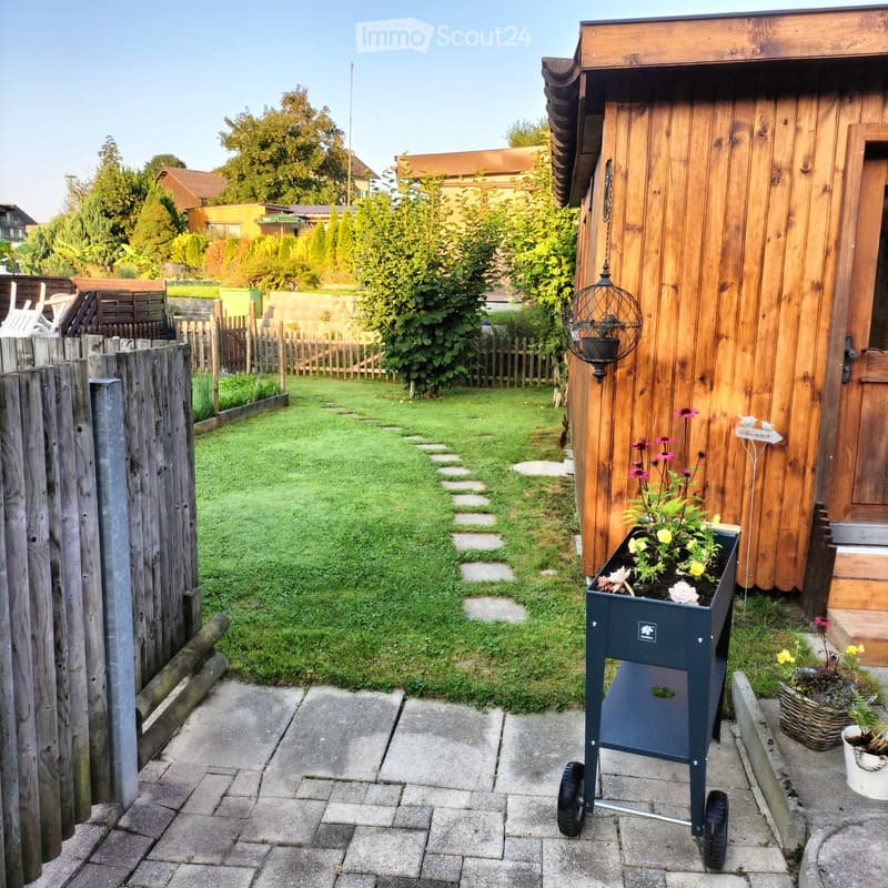 brick sidewalk, wooden fence, pathway of bricks leading to garden, green lawn, flowers in pot, metal stand with flower pot
