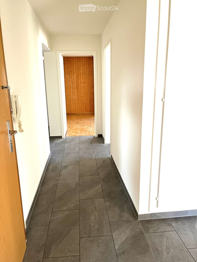 An empty hallway with tiled flooring, white walls, an open door leading to a wooden-paneled room, a wall-mounted phone, and a closed door on the right side.