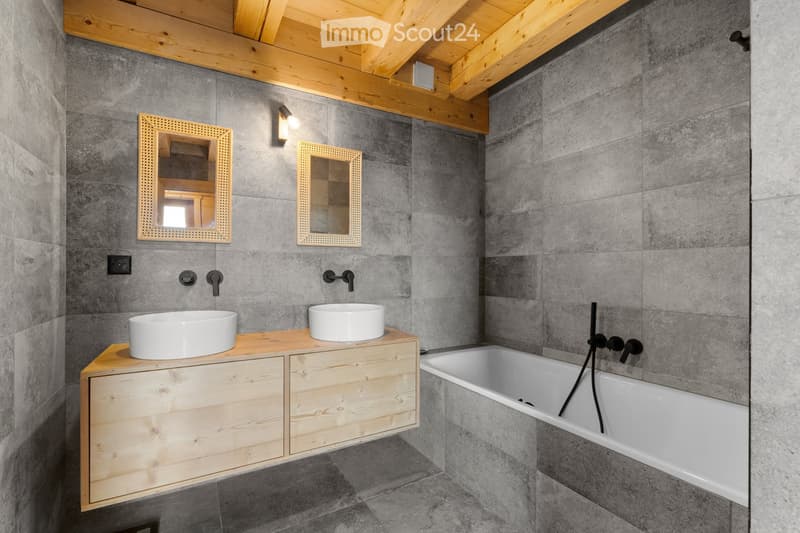 Spacious bathroom with gray tiled walls, wooden vanity with double sinks, framed mirrors, and a bathtub