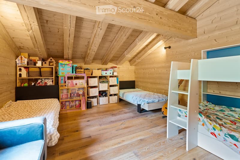 Spacious attic bedroom with wooden beamed ceiling, wooden floors, and various storage shelves and furniture for toys and personal items. The room includes a single bed and a bunk bed, as well as a seating area.