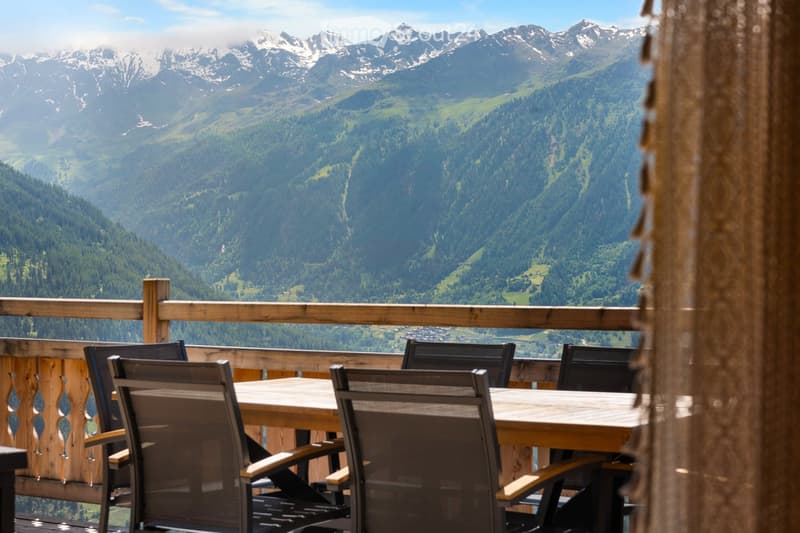 Wooden table and chairs on a balcony or deck overlooking a scenic mountain landscape with snow-capped peaks and lush green forests.