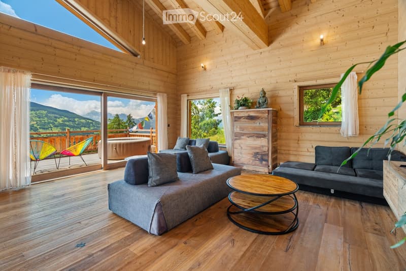 Spacious living room with wooden walls and ceiling, large windows overlooking a scenic mountain view, comfortable grey sofas, a wooden coffee table, and a wooden dresser with a Buddha statue on top