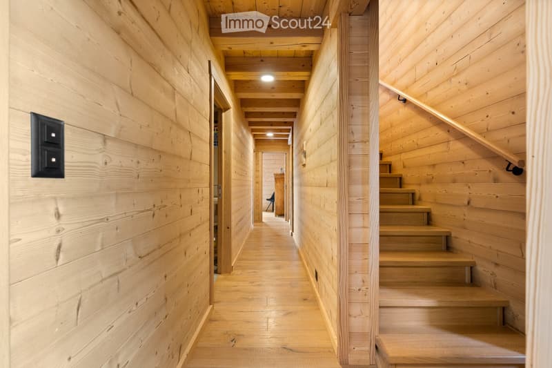 The image shows the interior of a wooden cabin or chalet. The walls and ceiling are made of natural wood panels, creating a warm and rustic atmosphere. The hallway features a wooden staircase leading to an upper level, and there are recessed lighting fixt