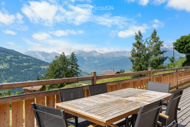 Wooden deck with a dining table and chairs, overlooking a scenic mountain landscape with forests and blue sky with clouds