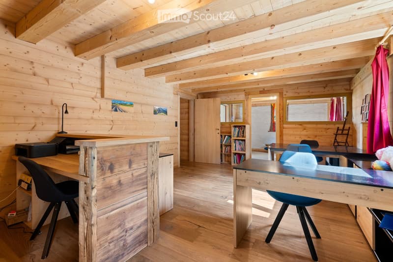 Cozy wooden cabin interior with exposed beams, wooden walls and floors, a desk with a computer, bookshelves, and a fireplace visible.