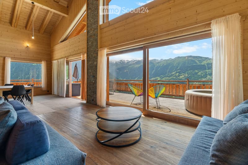 Spacious living room with wooden walls and ceiling, large windows offering a scenic mountain view, colorful chairs on the balcony, and a modern coffee table in the center of the room