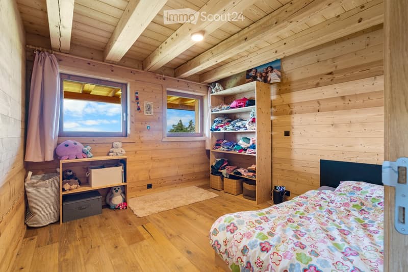 Cozy wooden bedroom with large windows, wooden ceiling beams, shelving unit with toys and storage, floral bedding, and a view of the outdoors