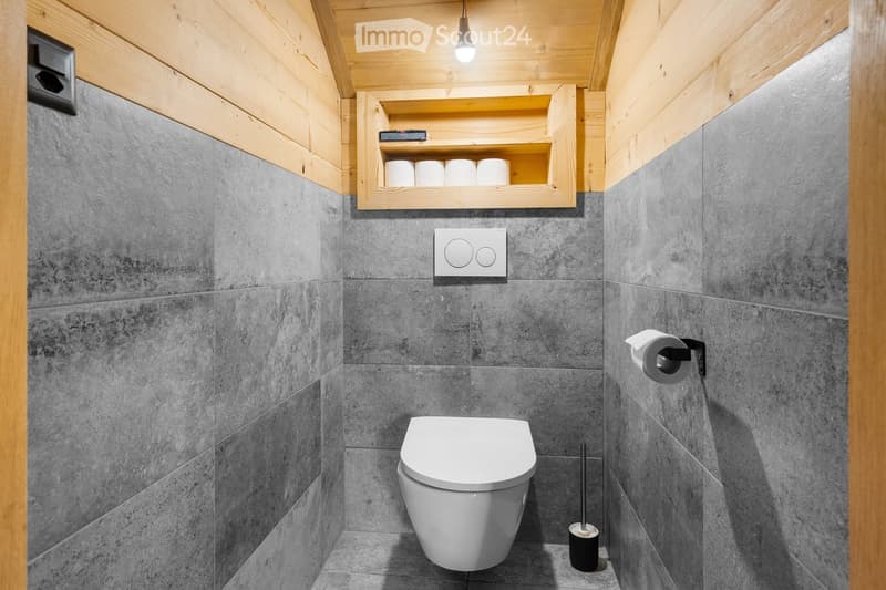 Bathroom with grey tile walls, wooden ceiling, toilet, toilet paper storage, and a light fixture.