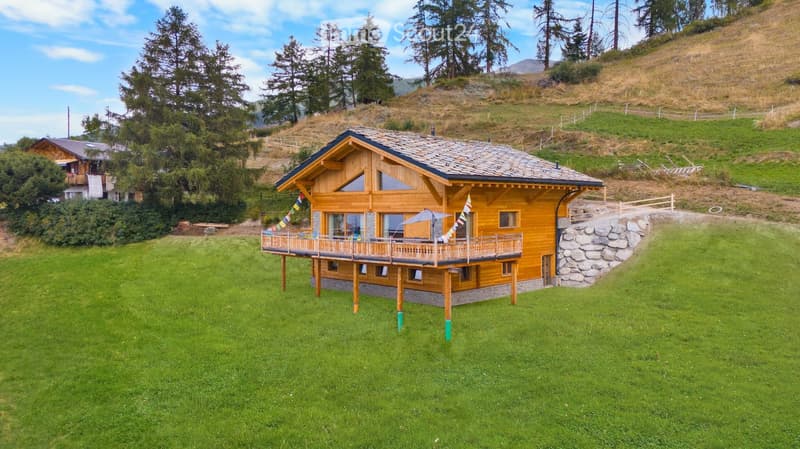 House with wooden exterior and tiled roof, elevated balcony, situated on grassy slope, stone walls for support