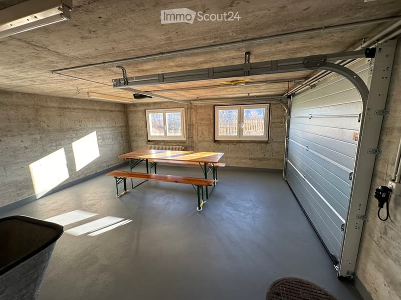 A room with concrete walls, floor and ceiling, large windows, a wooden bench table and two benches, an open garage door, a telephone outlet, a trash bin.