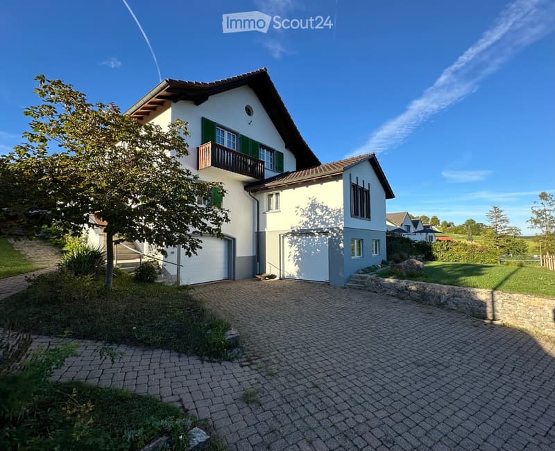 Two-story house with white exterior, green shutters, garage, and balcony on the upper level. The house is surrounded by a well-maintained garden and driveway.
