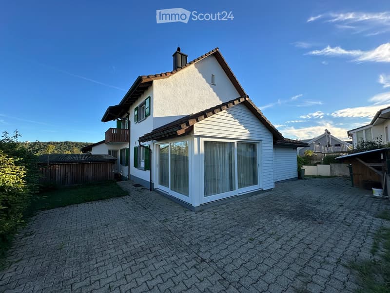 Two-story house with white exterior, green shutters, and a balcony on the upper level. The house features a pitched roof with wooden trim and is situated on a paved driveway with surrounding greenery.