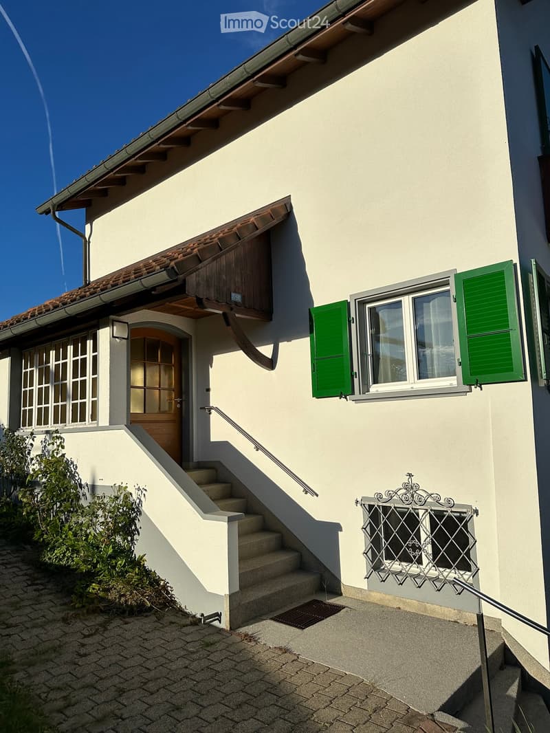 Two-story house with light beige walls, green shutters, and a tiled roof. Features a wooden front door, a staircase leading to the entrance, and a decorative metal railing on the balcony.