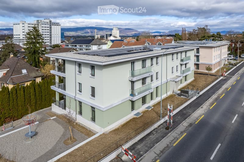 A modern residential building with multiple balconies, solar panels on the roof, and a street view