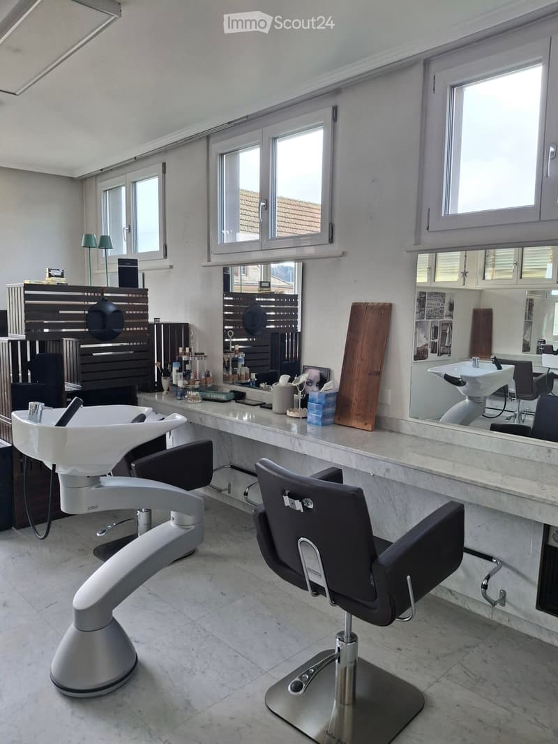 A white bathroom with white marble counter top and tiled floor, two white basins with faucets, mirror and three mirrors on the wall, salon chair in front of the counter, lamp and shelves with items