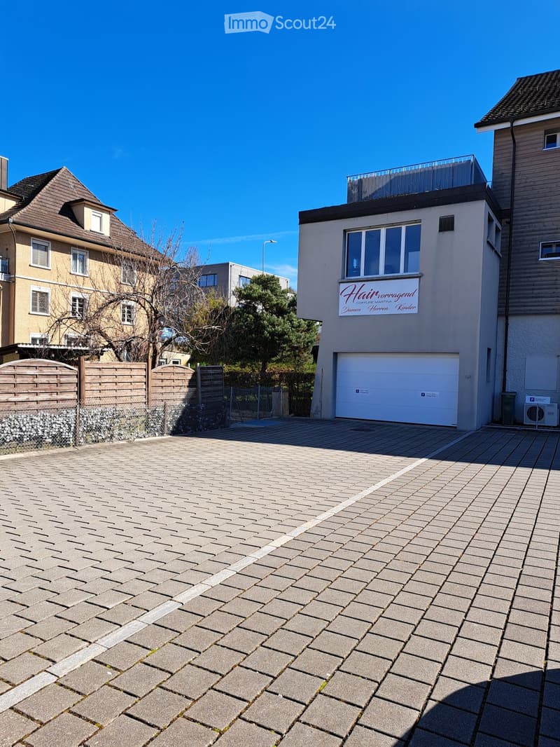 Front parking lot with a brick floor, wooden fence with a gate, white garage with windows, and two other neighboring buildings