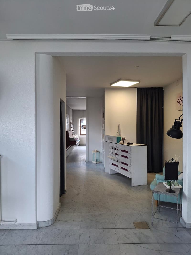 Open space with minimalist design, white walls, gray tile floor, large desk with computer, blue couch with table, shelves with storage boxes, and a lamp