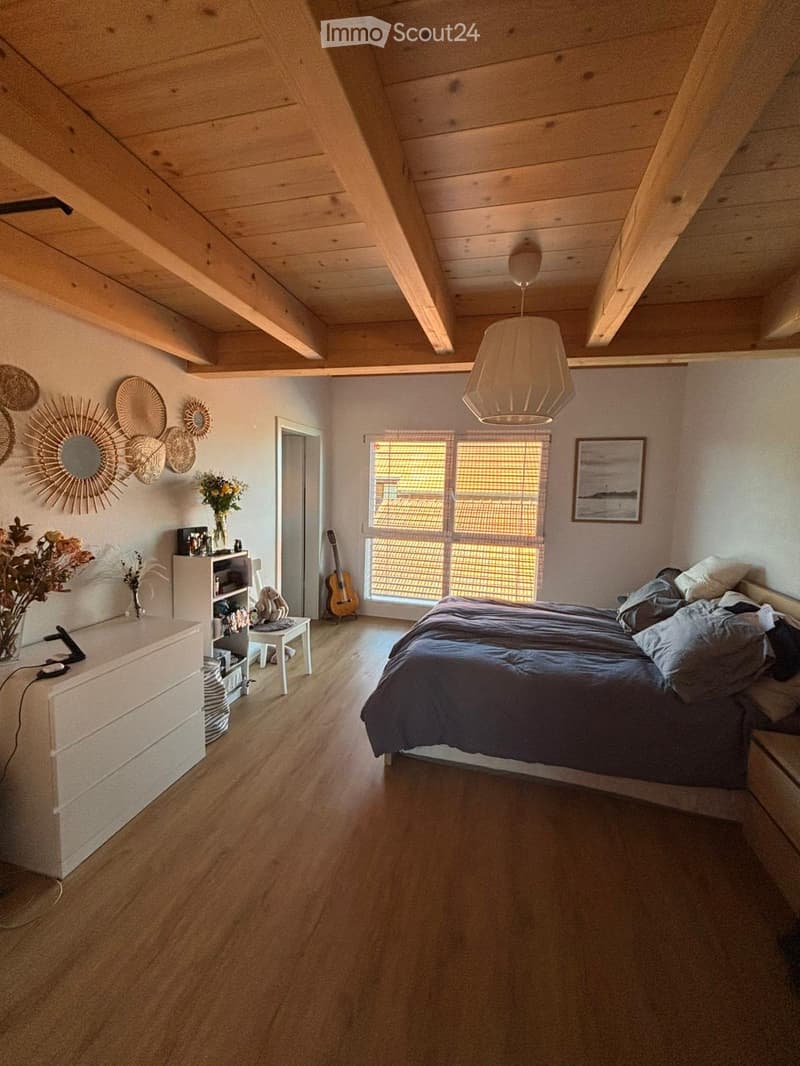 Bedroom with wooden floor, white walls, wooden ceiling beams, large window, bed, nightstand, cabinet, guitar, and decorative items.