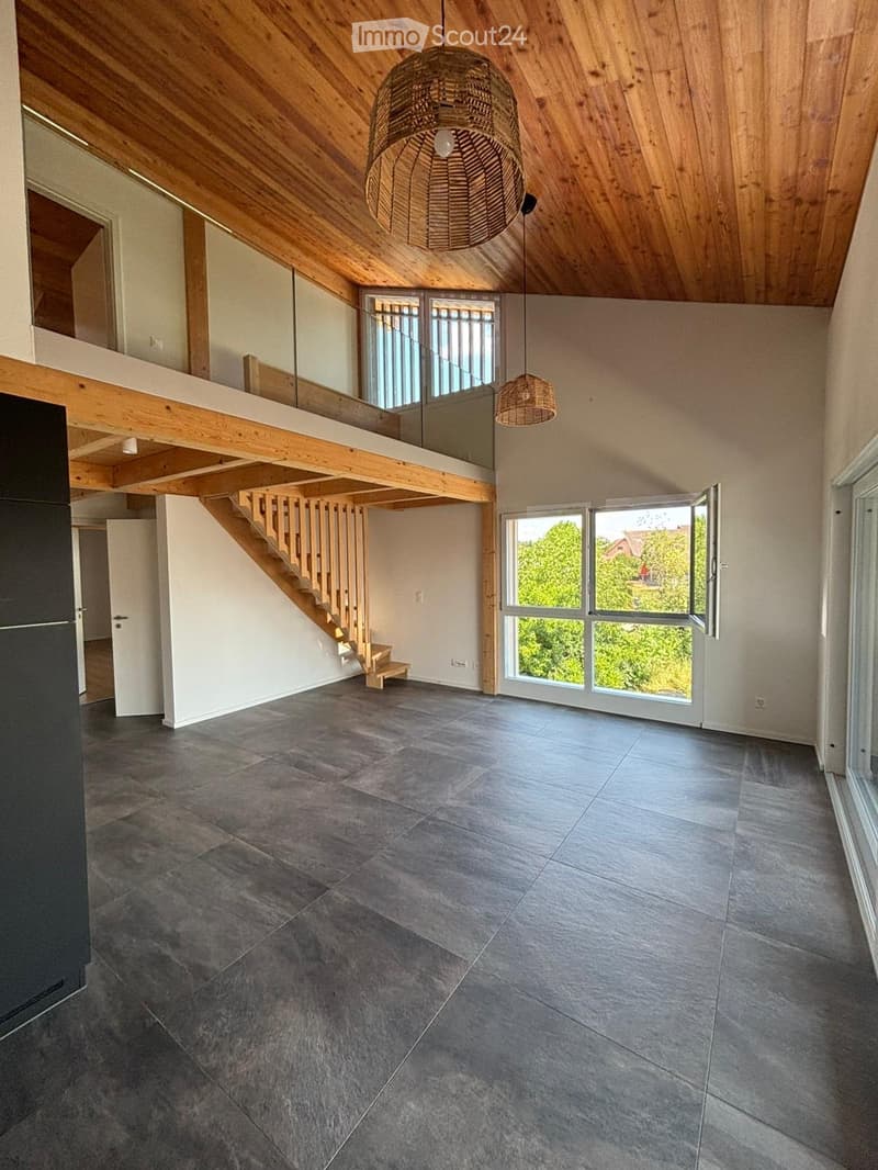 Open-plan living space with exposed wooden beams, grey tiled floor, large windows, and a wooden staircase leading to an upper level.