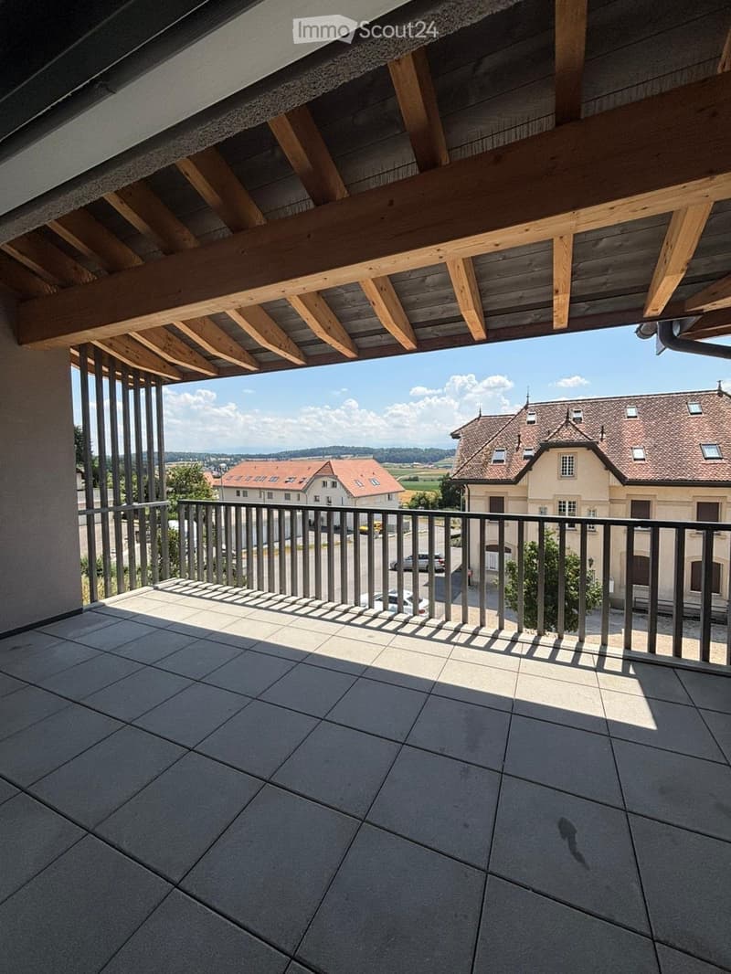 Balcony with tiled floor, metal railing, and wooden ceiling beams, overlooking a scenic countryside view with houses and fields.