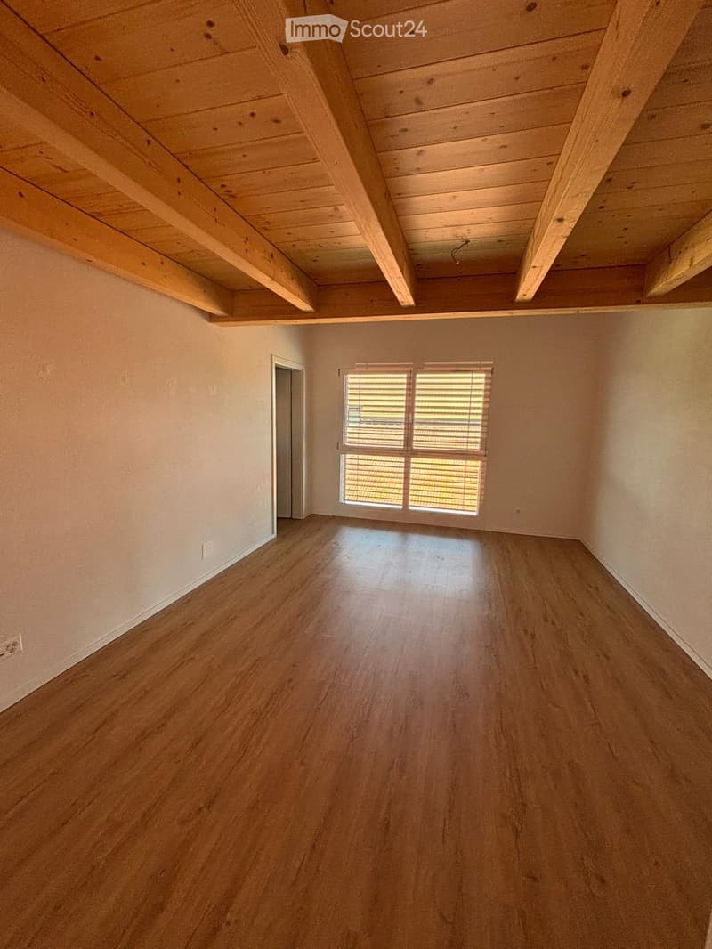 Empty room with wooden flooring, exposed wooden beams on the ceiling, and a window with blinds.