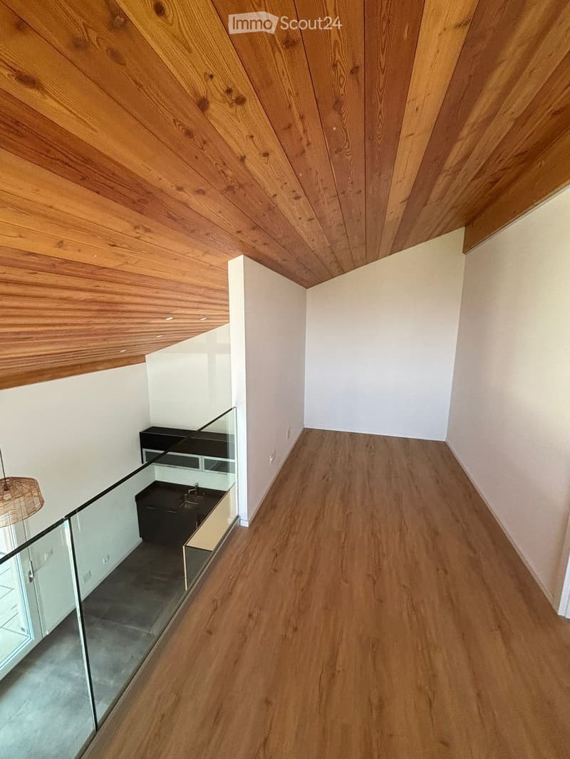 Modern hallway with wooden flooring and slanted wooden ceiling, featuring a glass railing leading to a small black vanity area with a sink and cabinets.