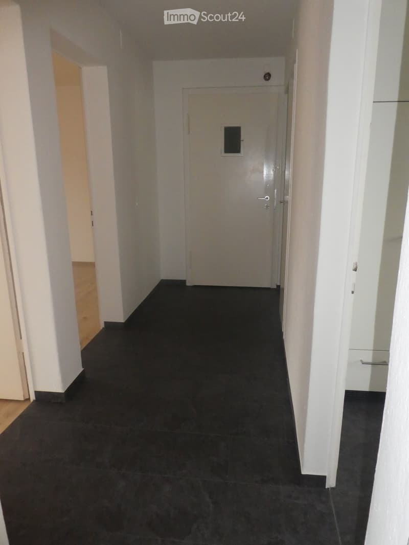 Corridor with white walls, black tiled floor, closed door, small glass window, small black stripe