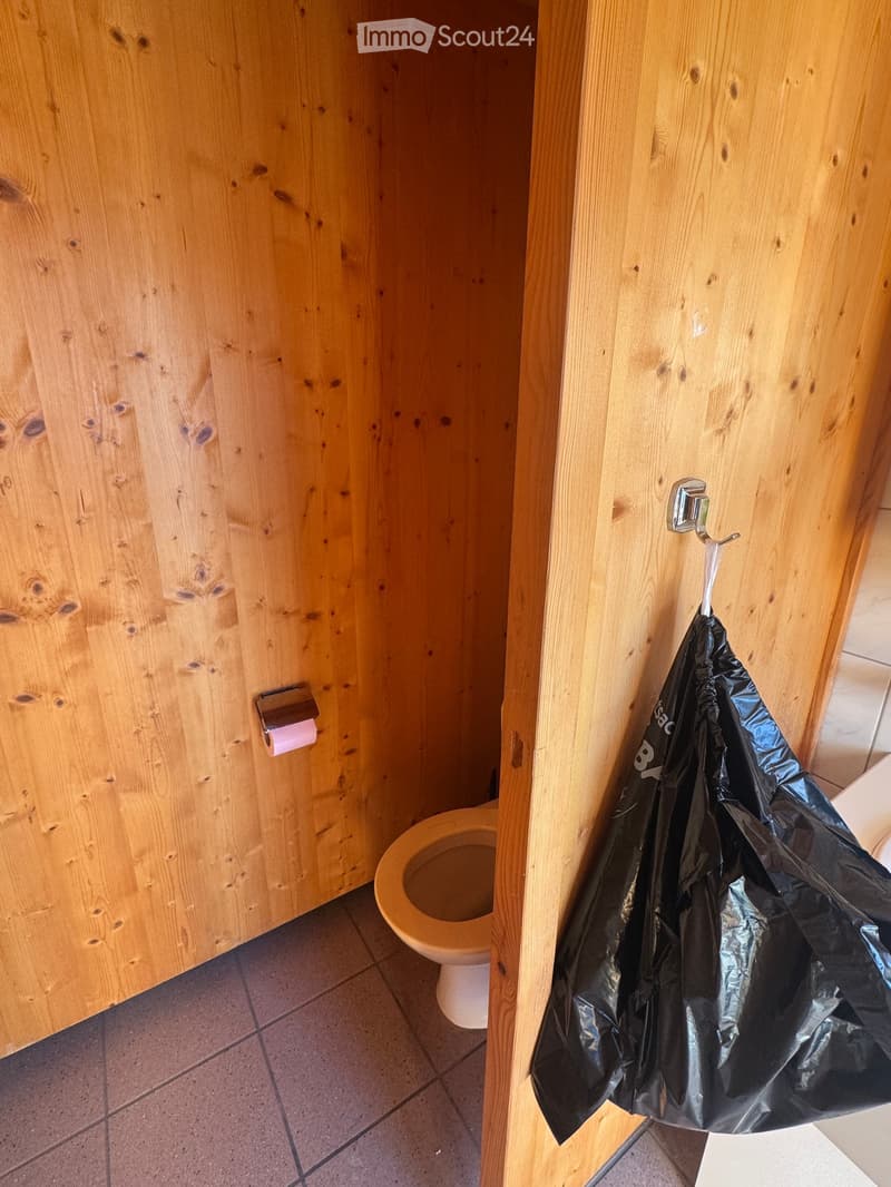Wooden paneled walls, tiled floor, toilet, roll of toilet paper, and a black plastic bag hanging on a hook.