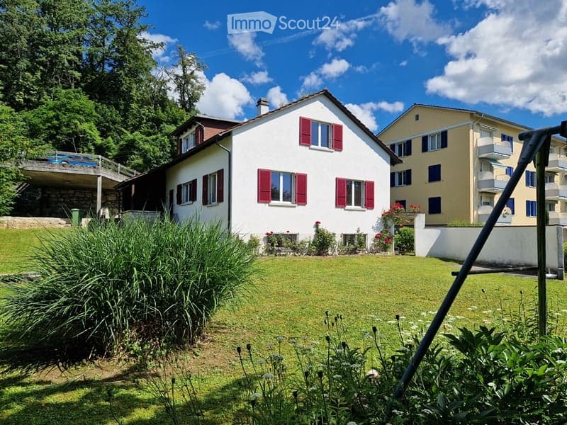 House with red shutters, surrounded by greenery and flowers, paved terrace, red flowers in front, white wall at back