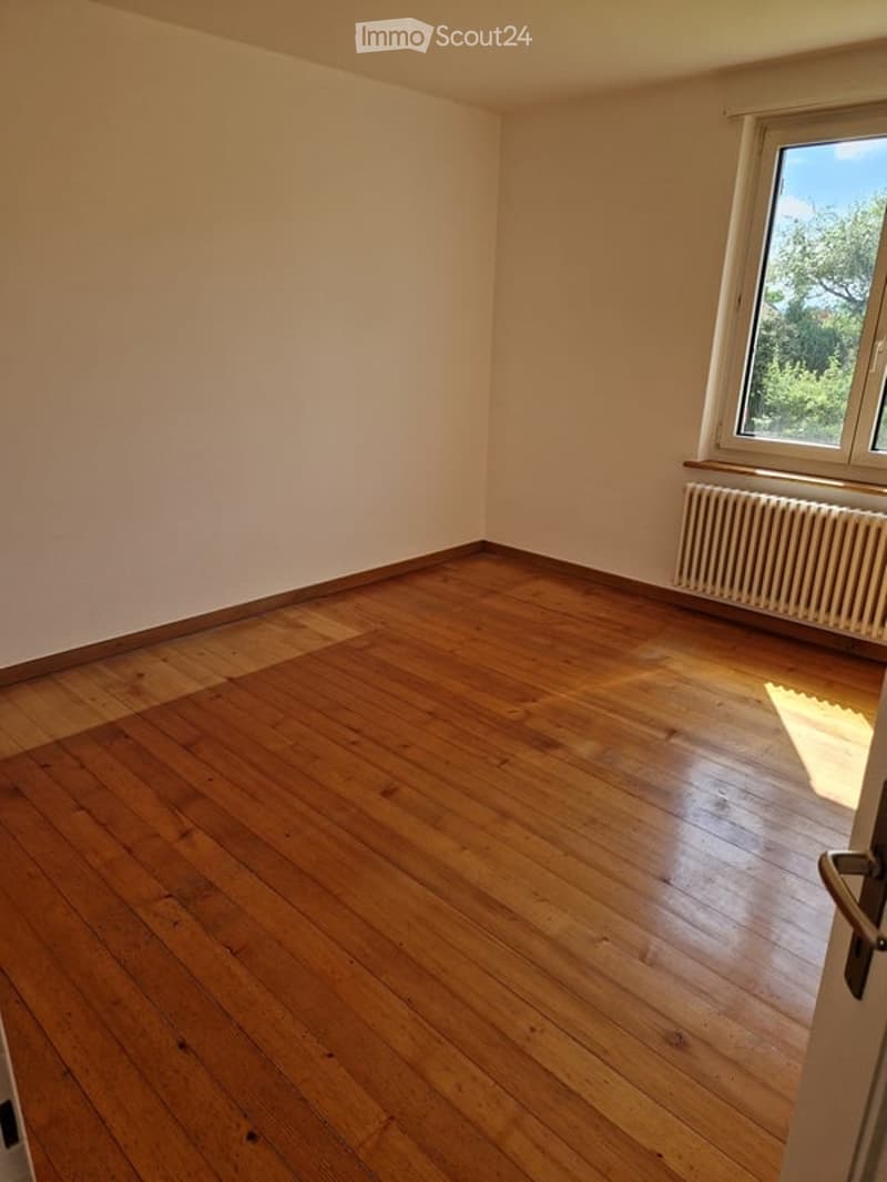 Empty room with hardwood floors, white walls, single window, and radiator. The room has an open door leading out.