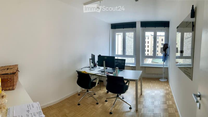 An empty office room with two black office chairs, a desk with computer, papers, and other items on it, and three windows overlooking outside.