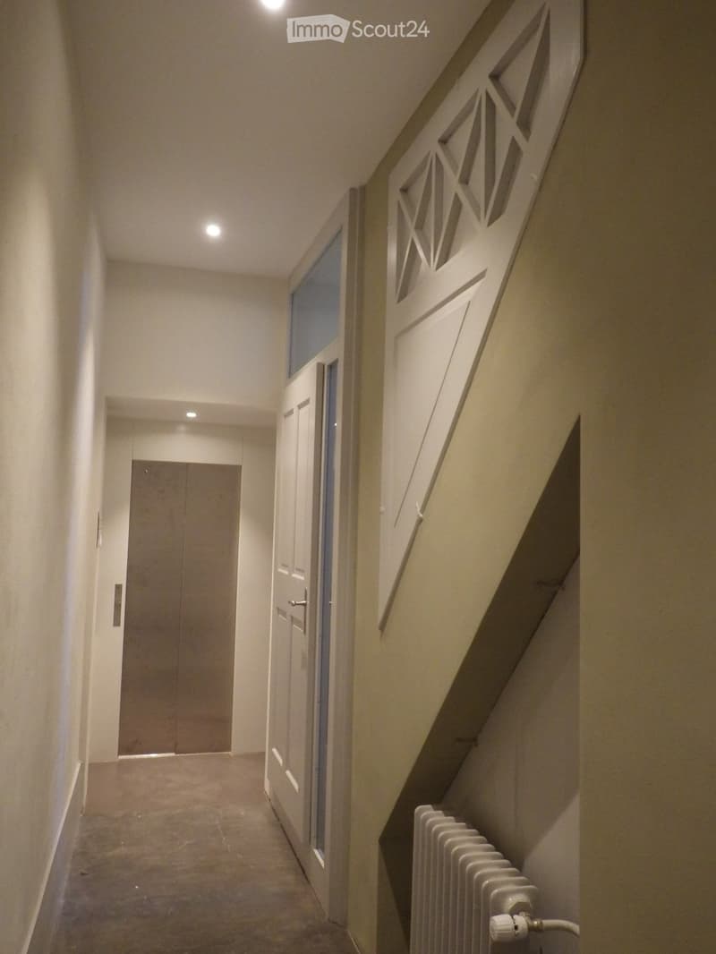 Corridor with white walls, gray floor, two doors, one with a glass window, a radiator, and a triangular design on the wall