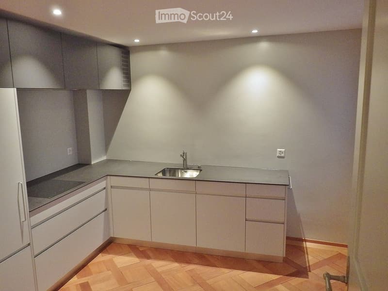Modern kitchen with white cabinets, wooden floor, gray countertop, stainless steel sink and faucet, electrical outlet, dishwasher, refrigerator.