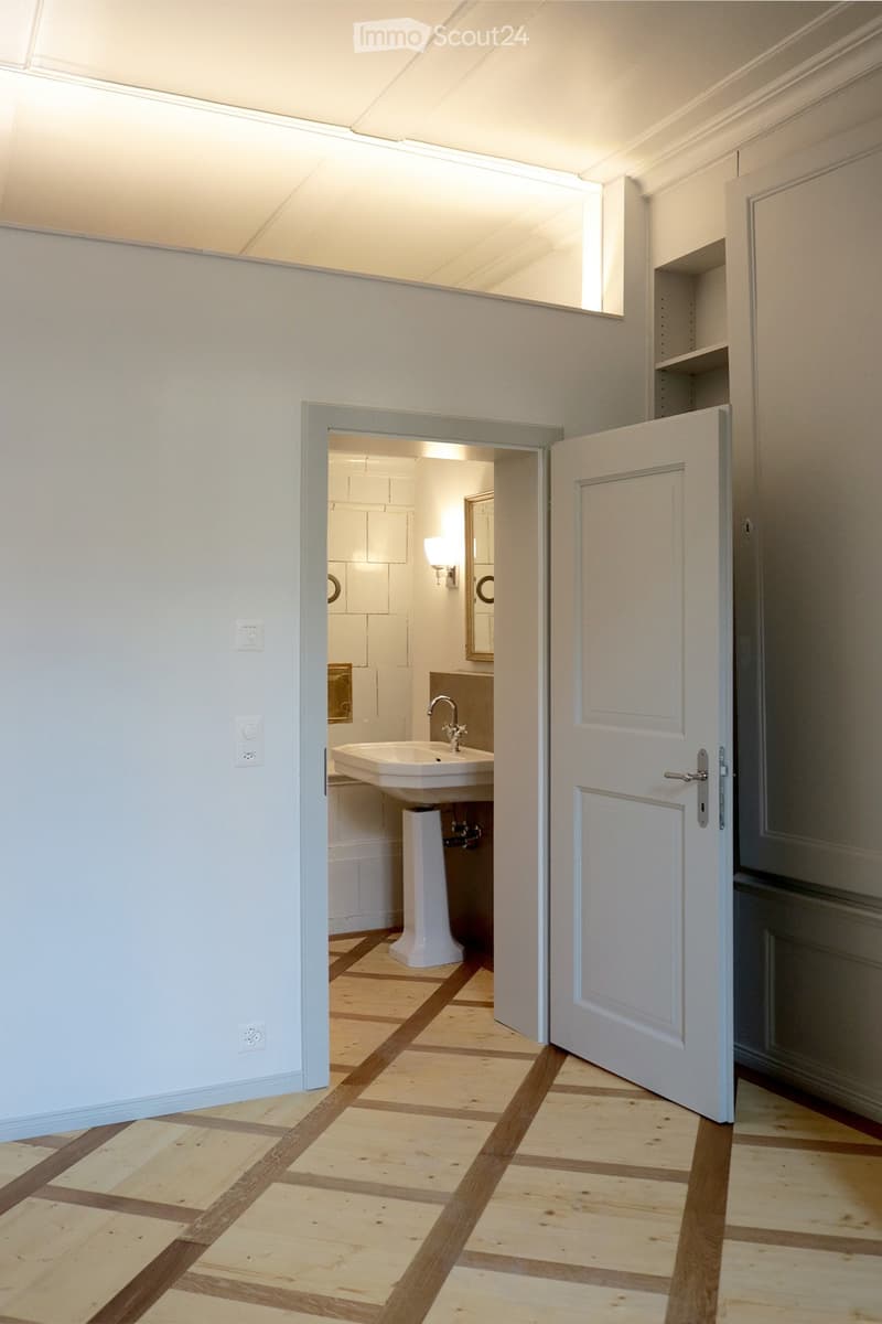Modern bathroom with wooden flooring, white walls, and a vanity with a sink. There's also a mirror mounted on the wall, and a window.