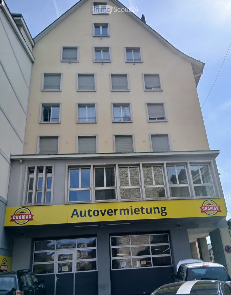 Four-story building with light yellow facade and multiple windows, featuring a yellow sign reading 'Autovermietung' and 'CHAMAS' above the entrance.