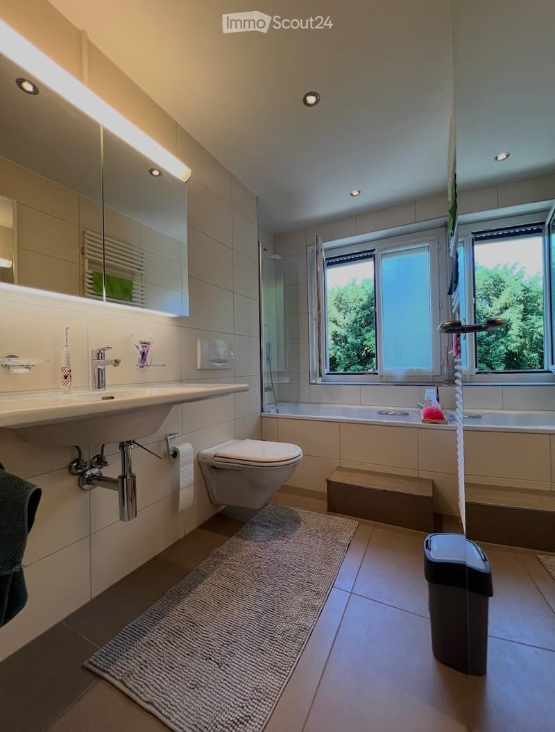 modern bathroom with a bathtub, sink, and toilet; large mirror; bright natural light from windows; tiled floor; rug
