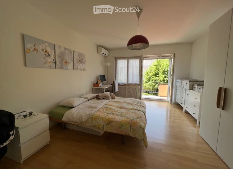 Double bed, wooden floor, white walls, large windows with balcony, white wardrobe, desk with computer, decorative paintings