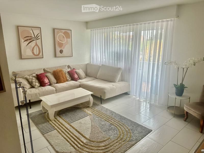Modern living room with a beige sectional sofa, colorful throw pillows, a white coffee table, a side table with a flower vase, a patterned rug, and two wall-mounted artworks.