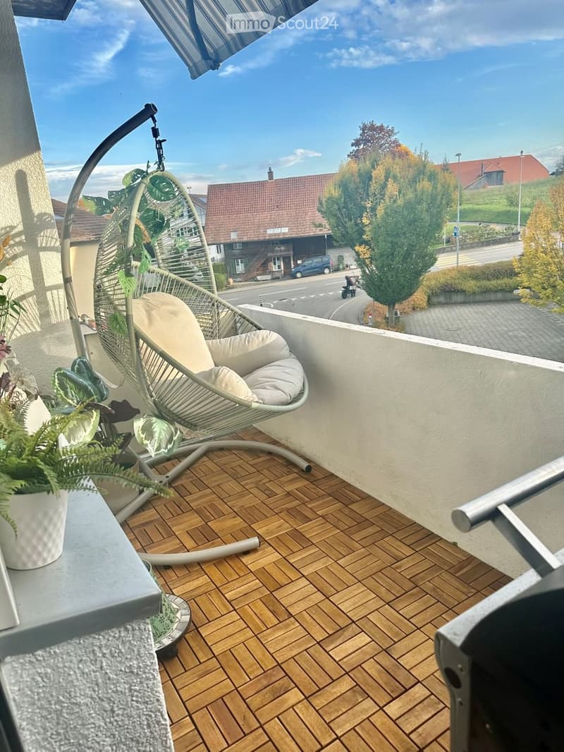 Wooden floored balcony with outdoor seating, hanging plant in pot and white cushion.