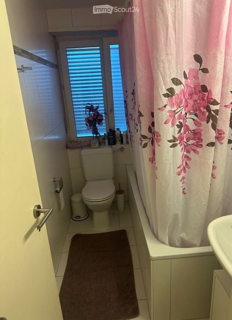 A bathroom with toilet, sink, bathtub, window, brown floor mat, curtains, and storage shelves.