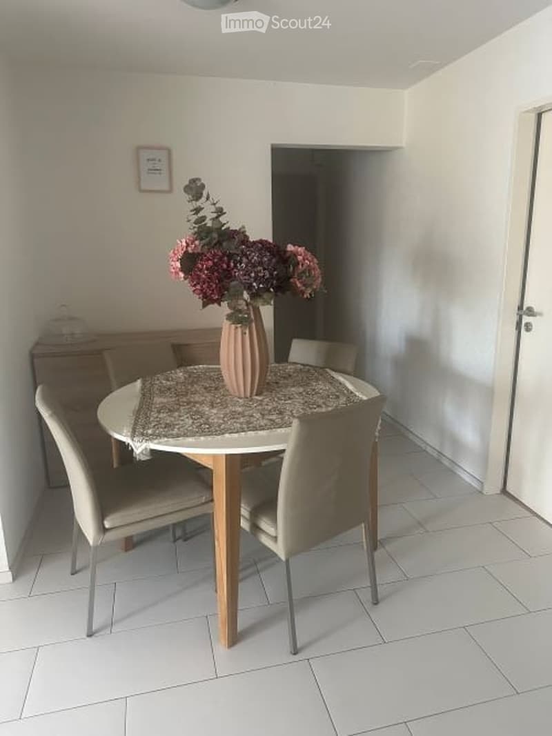 A table with a flower vase, surrounded by chairs. The room has white walls and tiled floor.