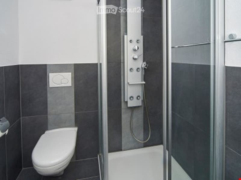 Modern bathroom with walk-in shower, white toilet, gray tiles, silver shower handle, and roll of toilet paper