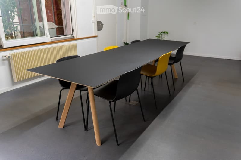 A modern, minimalist room with a long black table and several chairs, featuring a grey carpeted floor and white walls.